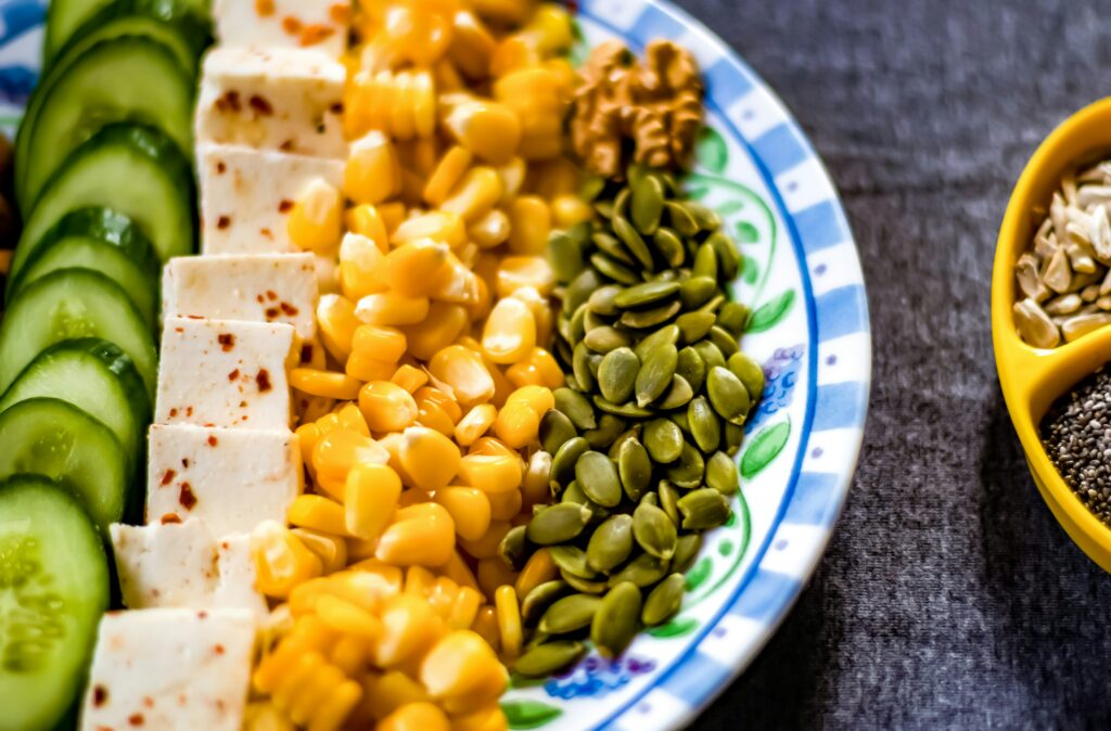 cucumber, tofu, pumpking seeds, and corn lined up on a blue and white plate